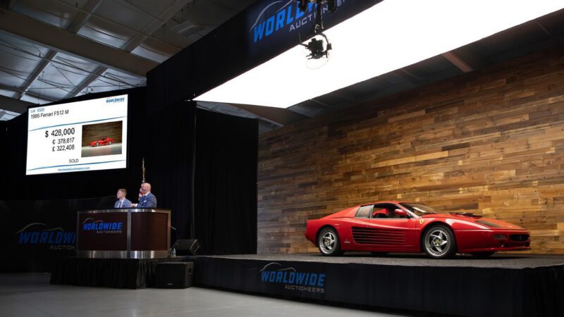 A red 1989 Ferrari is displayed on stage at a Worldwide Auctioneers Enthusiast Auction, with auctioneers at the podium and a screen showing the $428,000 sale price during the Spring Sales event.