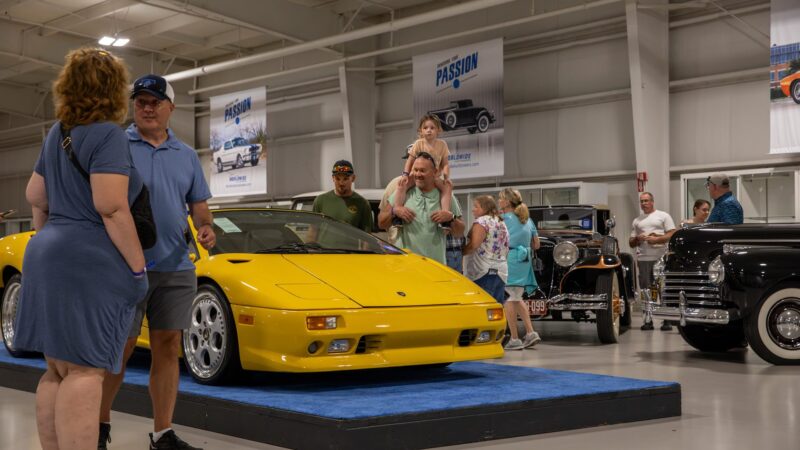 People gather around a yellow sports car on display at the Worldwide Auctioneers Enthusiast Auction, with vintage cars and informational posters visible in the background.