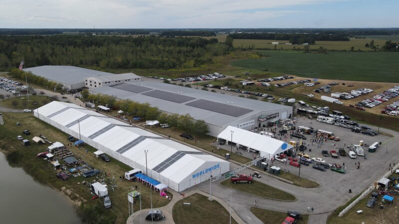 Aerial view of the Spring Sales event by Worldwide Auctioneers, featuring large tents, classic cars, and people outside a building with solar panels; parking lots and fields surround the venue at this Enthusiast Auction.