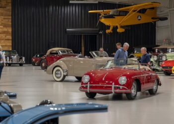 A group of people observe classic cars displayed in a spacious indoor showroom at the Worldwide Auctioneers Enthusiast Auction, with a yellow vintage airplane suspended above.