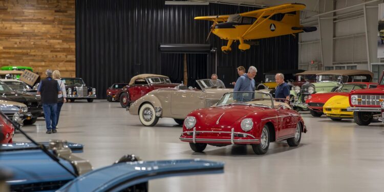 A group of people observe classic cars displayed in a spacious indoor showroom at the Worldwide Auctioneers Enthusiast Auction, with a yellow vintage airplane suspended above.