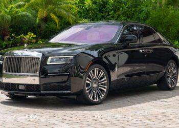 A 2024 Rolls-Royce Ghost collector’s car sedan parked on a stone driveway, surrounded by green foliage and palm trees.