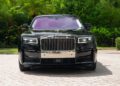 A 2024 Rolls-Royce Ghost is parked on a stone driveway, facing forward, with lush greenery and trees in the background—a true collector’s car.