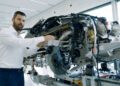 A man in a white shirt stands next to a partially assembled Bugatti, pointing at Tourbillon components in a bright, modern workshop.