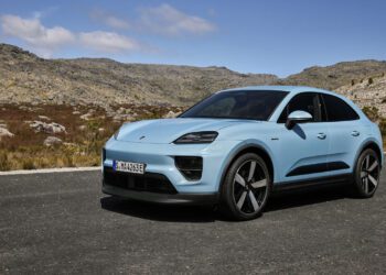 A light blue electric Macan SUV from Porsche Cars North America is parked on a paved road with rocky hills and a partly cloudy sky in the background.