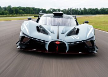 A grey Bugatti Bolide supercar drives on a racetrack, viewed from the front with trees and grass in the background.