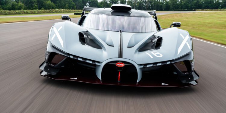 A grey Bugatti Bolide supercar drives on a racetrack, viewed from the front with trees and grass in the background.