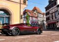 A maroon Bugatti W16 Mistral is parked on a cobblestone street in front of historic half-timbered buildings in the charming town square of Molsheim.