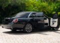 A 2024 Rolls-Royce Ghost luxury sedan with both rear doors open is parked on a stone driveway surrounded by green plants and trees—a true collector’s dream.