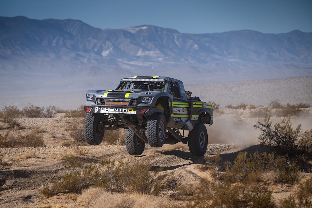 A lifted Brenthel Industries race truck leaps over desert terrain, kicking up dust in an epic off-road adventure, with mountains rising in the background.