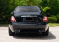 A black 2024 Rolls-Royce Ghost luxury sedan is parked on a paved driveway, viewed from the rear, with trees and greenery in the background—a true collector’s car.