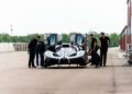 A group of people, including an expert support team, stand around a silver Bugatti Bolide with gullwing doors open on a race track, inspecting and preparing the exclusive vehicle.