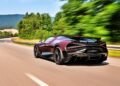 A red Bugatti W16 Mistral drives at high speed on a tree-lined road near Molsheim, with blurred scenery and hills in the background.