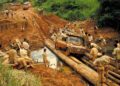 A muddy off-road vehicle, reminiscent of a Land Rover Classic to Showcase 10 Historic Expedition Icons at 2025 Pebble Beach Concours d'Elegance, is guided across a makeshift log bridge by a group in a rural, forested area. Other vehicles and people are nearby.