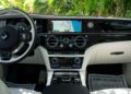 Luxury car dashboard featuring a digital display with navigation, climate controls, and "GHOST" branding—perfectly capturing the elegance of the 2024 Rolls-Royce Ghost, with lush greenery visible through the windshield.