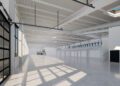 Spacious, empty industrial warehouse with high ceilings, large windows, exposed beams, and a mezzanine level along one wall—ideal as a Bugatti production facility. Sunlight casts shadows on the polished concrete floor.