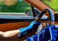 Person wearing blue racing gloves driving a Bugatti W16 Mistral, surrounded by tan leather interior, with blurred green scenery rushing past the window—a thrilling test drive near Molsheim.