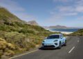A light blue Macan drives along a winding coastal road surrounded by green hills, with mountains and the ocean in the background under a partly cloudy sky—a stunning scene from Porsche Cars North America’s H1 2025 showcase.