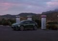 A green electric Macan SUV, part of the H1 2025 Porsche Cars lineup, is parked at a charging station in an empty lot, with mountains and a cloudy sky in the background.