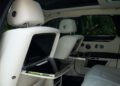 The image shows the rear interior of a 2024 Rolls-Royce Ghost with white leather seats, two screens on the seatbacks, and fold-out tray tables—a true collector’s car.