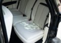 The image shows the rear seats of a 2024 Rolls-Royce Ghost with white upholstery, black seatbelts, and red seatbelt buckles. The car door is open, revealing part of the luxurious interior—a true collector’s car.