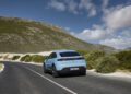 A light blue Porsche Taycan drives on a curving road through a scenic, mountainous landscape under a partly cloudy sky, reflecting the innovation seen in Porsche Cars North America as it looks ahead to H1 2025 growth.