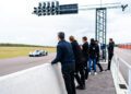 A group of people stand by a concrete barrier watching the exclusive Bugatti Bolide speed past on the track near a starting light structure.