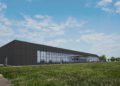 A modern, low-profile production facility with large windows and a dark facade sits on a grassy field under a partly cloudy sky.