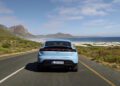 A light blue Porsche Taycan, part of the Porsche Cars North America lineup, drives on a coastal road with mountains on the left and the ocean on the right under a partly cloudy sky, highlighting H1 2025 Growth alongside models like the Macan.