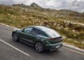 A green Porsche Taycan Cross Turismo, a highlight from Porsche Cars North America, drives on a paved road through a rocky countryside landscape under partly cloudy skies.