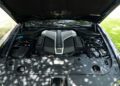 A car engine with a V12 badge is visible under an open hood, surrounded by clean engine components and greenery in the background—a true collector's car, reminiscent of the refined 2024 Rolls-Royce Ghost.