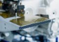 Close-up of a metallic honeycomb structure with a gold-colored surface, packaged within a mechanical assembly reminiscent of Bugatti engineering, with visible wiring and components in the background.