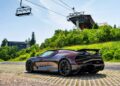 A dark-colored Bugatti W16 Mistral sports car is parked on a paved area near grassy hills in Molsheim, with a ski lift and buildings in the background under a clear sky, ready for its next test.