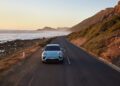 A light blue Macan drives along a coastal road at sunset, with mountains on one side and the ocean on the other—a stunning showcase of Porsche Cars, hinting at what’s to come in H1 2025.