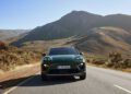A green Porsche Macan, one of the latest Porsche Cars for H1 2025, cruises along a paved road through a mountainous landscape under a clear blue sky.