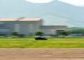 A Bugatti W16 Mistral speeds along a runway in Molsheim, with a blurred hangar and windsock in the background, indicating motion.