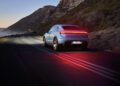 A white Porsche Macan from Porsche Cars North America drives along a coastal road at dusk, rear lights aglow and mountains in the background—anticipating the innovations of H1 2025.