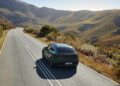 A green Porsche Macan drives along a winding, empty road through hilly, sunlit countryside under a clear sky, showcasing the elegance of Porsche Cars in H1 2025.