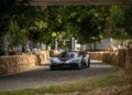 A black and white camouflaged Zenvo Aurora sports car drives on a track lined with hay bales at a motorsport event, making its global debut, with a “START” sign visible in the background.