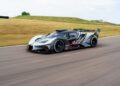 An exclusive black and silver Bugatti Bolide race car speeds along a paved track, surrounded by grassy fields in the background.