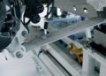 Close-up of a mechanical arm or suspension component, similar to those in Bugatti Tourbillon engineering, attached to a metal structure with visible wires and connectors in an industrial setting.