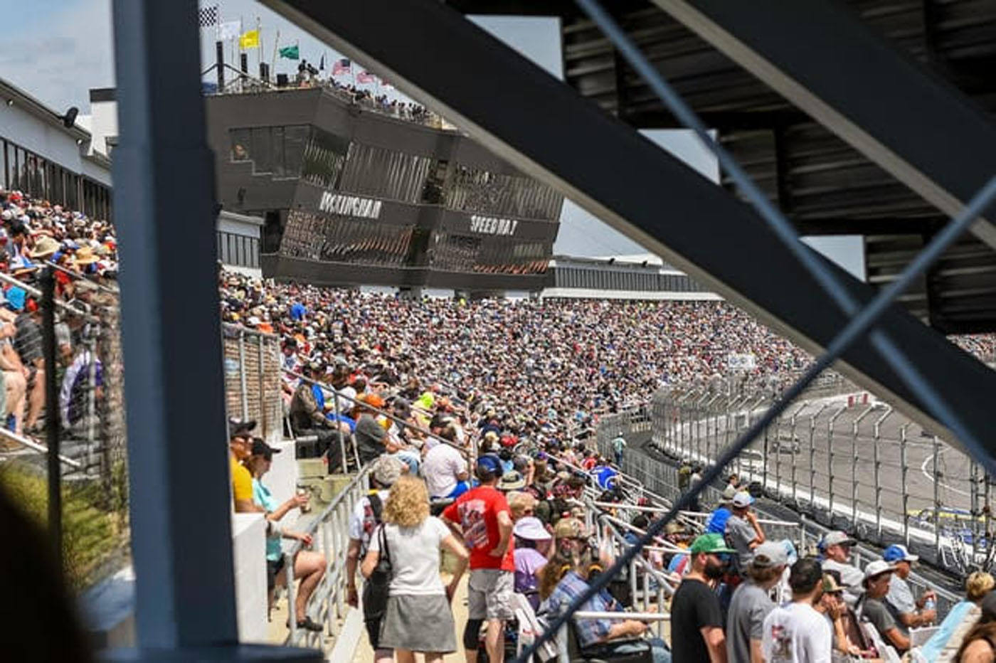 A large crowd of spectators fills the bleachers at Rockingham Speedway, the ultimate playground for racing fans, with the track and infield visible under a sunny sky.
