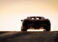 A rear view of a black Bugatti Tourbillon sports car on a road at sunset, its taillights illuminated and the sun creating a bright silhouette effect.
