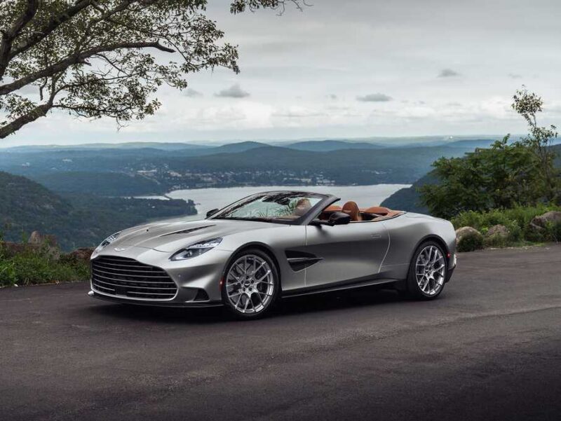 A 2026 Aston Martin Vanquish V12 convertible sports car parked on a paved road, offering a stunning view of hills, trees, and a river in the background.