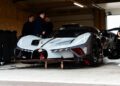 An exclusive Bugatti Bolide track car sits in a garage as an expert support team prepares the vehicle for use, with several people standing nearby.