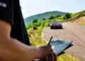 A person writes on a clipboard in the foreground while a Bugatti W16 Mistral, likely out for a test, is parked on a rural road with green hills reminiscent of Molsheim in the background.