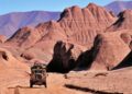 A rugged Land Rover Classic drives along a dirt road through a rocky desert landscape with large reddish-brown hills and distant mountains, echoing the spirit of the 10 historic expedition icons to be showcased at the 2025 Pebble Beach Concours d'Elegance.