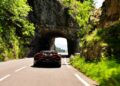 A sleek Bugatti W16 Mistral drives through a tunnel carved into a rocky cliff on a scenic, winding road near Molsheim, surrounded by greenery during a thrilling test run.