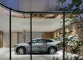 A silver car is parked inside the World's First Maybach Brand Center, a modern, glass-walled building with wood accents in Seoul, viewed from an outdoor garden area at dusk.