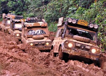 Three mud-covered Land Rover Defenders, true historic expedition icons, drive in a line through a narrow, muddy off-road track surrounded by dense green vegetation.